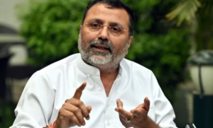 This image shows Nishikant Dubey, a Member of Parliament from the Bharatiya Janata Party (BJP), standing indoors. He is dressed in a simple white long-sleeved shirt, has a short salt-and-pepper beard, and is wearing spectacles. He is looking slightly away from the camera with a neutral, contemplative expression. This image is often used in the context of his recent February 2026 announcement to establish a library documenting the history of the Nehru-Gandhi family.
