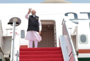This image shows Indian Prime Minister Narendra Modi waving while standing at the top of the stairs of an aircraft. He is dressed in a traditional white kurta-pyjama paired with a dark sleeveless jacket. He is positioned at the doorway of the plane, which features the State Emblem of India near the door. A red carpet covers the boarding stairs, and the scene suggests he is either departing for or arriving from an official visit.