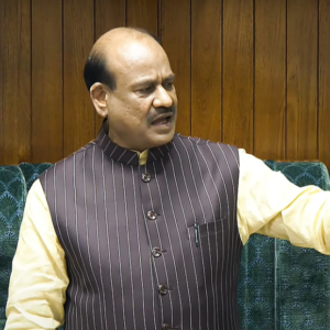 लोकसभा अध्यक्ष ओम बिरला की एक क्लोज-अप तस्वीर, जिसमें वे हल्के पीले रंग के कुर्ते और गहरे रंग की धारीदार नेहरू जैकेट पहने हुए हैं। वे लकड़ी के पैनल वाली पृष्ठभूमि के सामने खड़े होकर कुछ बोल रहे हैं।