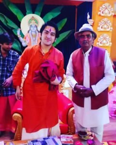 धीरेंद्र कृष्ण शास्त्री नारंगी रंग के कुर्ते में मुस्कुराते हुए खड़े हैं। उनके साथ एक अन्य व्यक्ति सफेद कुर्ता-पायजामा, नेहरू जैकेट और टोपी पहने खड़ा है। पृष्ठभूमि में भगवान कृष्ण की एक तस्वीर और सजावटी दीवार दिखाई दे रही है।