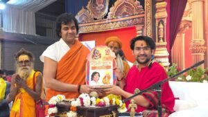 एक मंच पर दो प्रमुख व्यक्ति खड़े हैं। बाईं ओर एक व्यक्ति भगवा वस्त्र में है और दाईं ओर धीरेंद्र कृष्ण शास्त्री (बागेश्वर धाम सरकार) लाल वस्त्र में हैं। दोनों मिलकर एक पोस्टर या निमंत्रण पत्र पकड़े हुए हैं जिस पर धार्मिक चित्र बने हैं। उनके पीछे अन्य साधु-संत भी दिखाई दे रहे हैं।