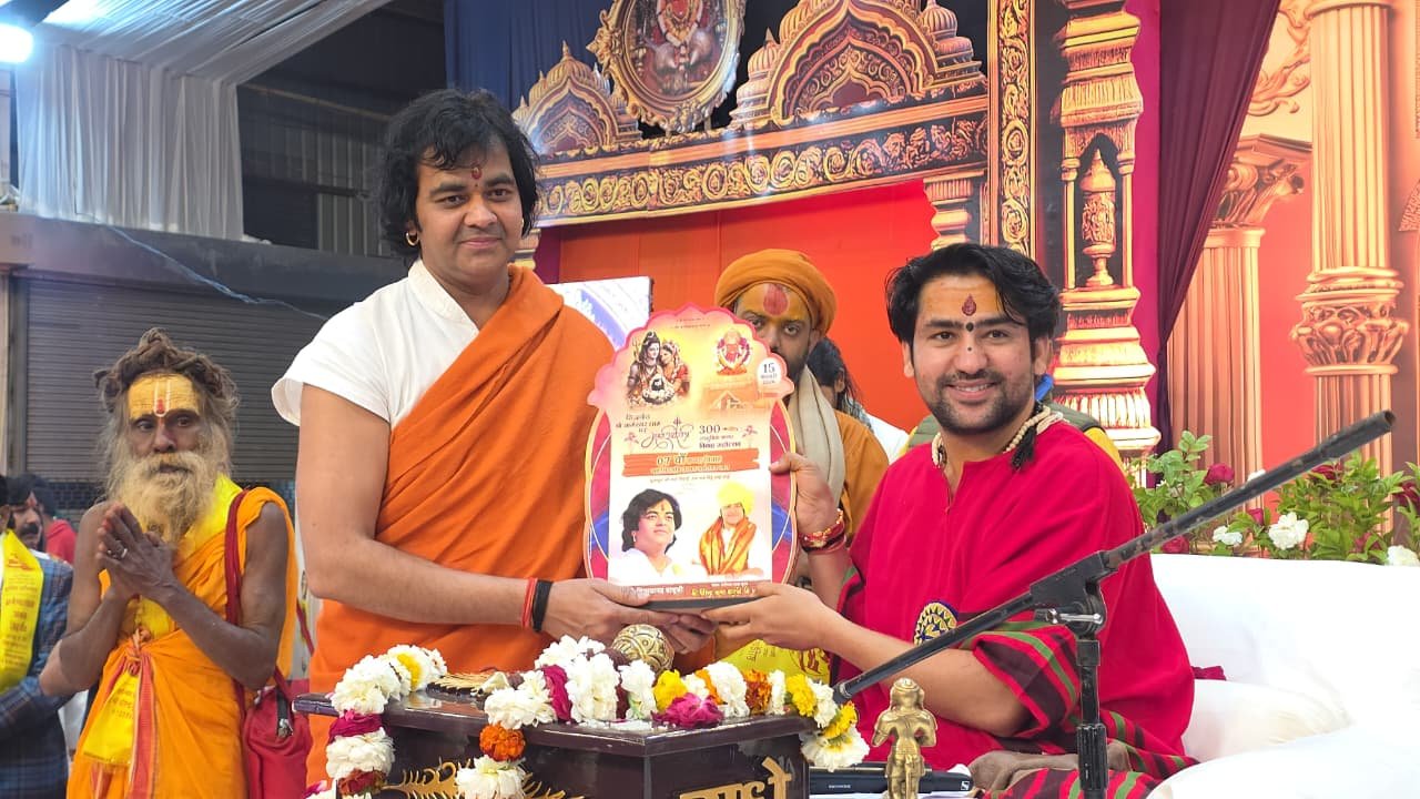 एक मंच पर दो प्रमुख व्यक्ति खड़े हैं। बाईं ओर एक व्यक्ति भगवा वस्त्र में है और दाईं ओर धीरेंद्र कृष्ण शास्त्री (बागेश्वर धाम सरकार) लाल वस्त्र में हैं। दोनों मिलकर एक पोस्टर या निमंत्रण पत्र पकड़े हुए हैं जिस पर धार्मिक चित्र बने हैं। उनके पीछे अन्य साधु-संत भी दिखाई दे रहे हैं।