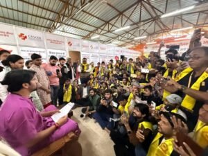 एक बड़े हॉल या पंडाल का दृश्य जहाँ धीरेंद्र कृष्ण शास्त्री एक ऊंची कुर्सी पर बैठकर कागज़ पढ़ रहे हैं। उनके सामने बड़ी संख्या में लोग ज़मीन पर बैठे हैं और अपने मोबाइल फोन से उनकी फोटो या वीडियो बना रहे हैं। दीवार पर 'कैंसर हीलर' के विज्ञापन बोर्ड लगे हैं।