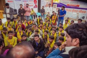 एक बड़े कमरे के अंदर का दृश्य जहाँ बहुत से लोग ज़मीन पर बैठे हैं। उनमें से अधिकांश ने पीले रंग के पटके (स्कार्फ) पहने हुए हैं। पृष्ठभूमि में 'कैंसर हीलर सेंटर' के पोस्टर और बैनर लगे हैं। कुछ लोग खड़े होकर भीड़ की तस्वीर खींच रहे हैं या वीडियो बना रहे हैं।