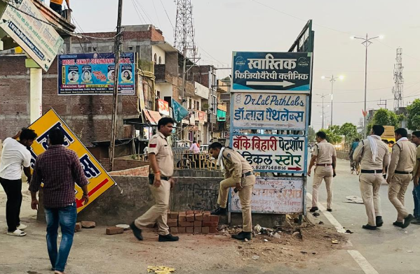 एक शहरी सड़क का दृश्य है जहाँ पुलिसकर्मी एक व्यावसायिक परिसर के बाहर खड़े हैं। सड़क के किनारे कई व्यावसायिक बोर्ड लगे हैं, जिन पर "स्वास्तिक फिजियोथेरेपी क्लिनिक", डॉ. लाल पैथलैब्स और "बाँके बिहारी पेटशॉप" जैसे नाम लिखे हुए हैं। खाकी वर्दी पहने चार-पांच पुलिस अधिकारी वहां मौजूद हैं, जिनमें से एक अधिकारी नीचे झुककर कुछ निरीक्षण कर रहा है। बाईं ओर, एक व्यक्ति पीले रंग का एक बड़ा बोर्ड हटाता हुआ दिखाई दे रहा है। पृष्ठभूमि में बिजली के ऊंचे टॉवर, बहुमंजिला इमारतें और अन्य दुकानें नजर आ रही हैं। यह दृश्य संभवतः नगर निगम या पुलिस द्वारा किए जा रहे अतिक्रमण हटाओ अभियान या किसी दुकान की जांच का है।