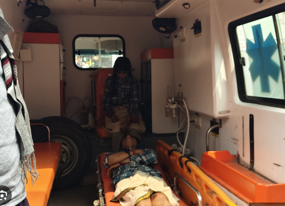 एक एम्बुलेंस के अंदर का दृश्य है, जहाँ एक घायल व्यक्ति स्ट्रेचर पर लेटा हुआ है। स्ट्रेचर के पास एक अन्य व्यक्ति बैठा है जो संभवतः घायल की देखभाल कर रहा है। एम्बुलेंस के अंदर चिकित्सा उपकरण, ऑक्सीजन सिलेंडर और नारंगी रंग की सीटें दिखाई दे रही हैं। खिड़की पर एम्बुलेंस का 'तारा' चिन्ह बना हुआ है। यह दृश्य किसी आपातकालीन चिकित्सा स्थिति या दुर्घटना के बाद अस्पताल ले जाने के दौरान का है।