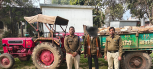एक खुले मैदान का दृश्य है जहाँ पुलिसकर्मियों ने अवैध परिवहन में लिप्त एक वाहन को पकड़ा है। बाईं ओर एक लाल रंग का महिंद्रा ट्रैक्टर खड़ा है, जिसके पीछे हरे रंग की ट्रॉली जुड़ी हुई है। ट्रॉली बड़े-बड़े पत्थरों (stones) से पूरी तरह भरी हुई है। ट्रैक्टर और ट्रॉली के बीच में खाकी वर्दी पहने दो पुलिस अधिकारी खड़े हैं, और उनके साथ एक अन्य व्यक्ति खड़ा है जिसका चेहरा धुंधला (blurred) कर दिया गया है। पृष्ठभूमि में एक सफेद रंग की छोटी इमारत और कुछ घने पेड़ दिखाई दे रहे हैं। यह दृश्य अवैध पत्थर उत्खनन या परिवहन के विरुद्ध की गई कानूनी कार्यवाही को दर्शाता है।