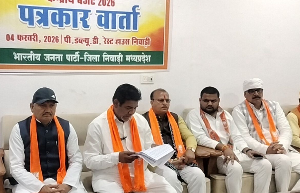 एक प्रेस कॉन्फ्रेंस का दृश्य है जहाँ भारतीय जनता पार्टी (BJP) के नेता एक सोफे पर बैठे हैं। पीछे एक बैनर लगा है जिस पर हिंदी में "पत्रकार वार्ता" और तारीख "04 फरवरी, 2026" लिखी हुई है। सोफे पर बैठे नेता भगवा रंग के पटके (scarves) पहने हुए हैं। केंद्र में बैठा व्यक्ति कुछ कागजात पढ़ रहा है, जबकि अन्य नेता गंभीरता से सुन रहे हैं। बैनर पर स्थान "पी.डब्ल्यू.डी. रेस्ट हाउस निवाड़ी" अंकित है।