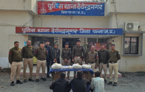 पन्ना जिले के देवेन्द्रनगर पुलिस थाने के सामने पुलिस अधिकारियों और पकड़े गए आरोपियों का एक दृश्य है। थाने की इमारत के बाहर पुलिस की एक टीम वर्दी में खड़ी है। उनके सामने एक नीली मेज पर सफेद रंग की बड़ी पोटलियाँ और पैकेट रखे हुए हैं, जो संभवतः जब्त किया गया मादक पदार्थ या अवैध सामग्री है। मेज के आगे जमीन पर तीन आरोपी पीठ करके बैठे हुए हैं। थाने के बोर्ड पर स्पष्ट रूप से "पुलिस थाना देवेन्द्रनगर, जिला पन्ना (म.प्र.) लिखा हुआ है।