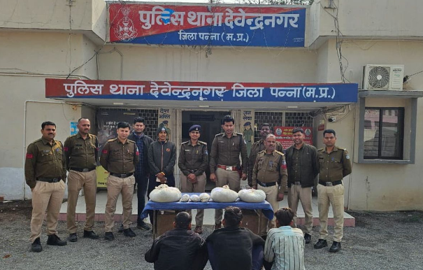 पन्ना जिले के देवेन्द्रनगर पुलिस थाने के सामने पुलिस अधिकारियों और पकड़े गए आरोपियों का एक दृश्य है। थाने की इमारत के बाहर पुलिस की एक टीम वर्दी में खड़ी है। उनके सामने एक नीली मेज पर सफेद रंग की बड़ी पोटलियाँ और पैकेट रखे हुए हैं, जो संभवतः जब्त किया गया मादक पदार्थ या अवैध सामग्री है। मेज के आगे जमीन पर तीन आरोपी पीठ करके बैठे हुए हैं। थाने के बोर्ड पर स्पष्ट रूप से "पुलिस थाना देवेन्द्रनगर, जिला पन्ना (म.प्र.) लिखा हुआ है।