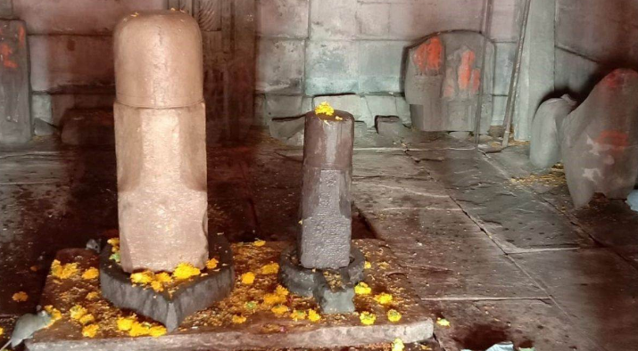 पत्थर से बने शिवलिंग का दृश्य, जिसमें एक बड़ा और एक छोटा शिवलिंग मंदिर के अंदर स्थापित हैं। शिवलिंगों पर गेंदा फूल चढ़ाए गए हैं और आसपास पत्थर का फर्श तथा मंदिर की दीवारें दिखाई दे रही हैं। यह एक प्राचीन मंदिर का आंतरिक दृश्य प्रतीत होता है