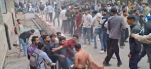 एक सार्वजनिक स्थान पर भीड़ और हंगामे का दृश्य है। चित्र के केंद्र में लोगों का एक बड़ा समूह दिखाई दे रहा है जो आपस में धक्का-मुक्की और विवाद करते हुए प्रतीत हो रहे हैं। कुछ युवक एक-दूसरे को पकड़ने और रोकने की कोशिश कर रहे हैं। अग्रभूमि में एक व्यक्ति बिना शर्ट के खड़ा है, जबकि अन्य लोग सामान्य कपड़ों में हैं। पृष्ठभूमि में बड़ी संख्या में छात्र या स्थानीय निवासी खड़े होकर इस स्थिति को देख रहे हैं। यह दृश्य किसी कॉलेज परिसर या सार्वजनिक स्थल पर हुए विवाद, विरोध प्रदर्शन या झड़प को दर्शाता है।