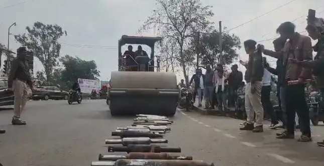 एक सड़क का दृश्य है जहाँ पुलिस की मौजूदगी में एक रोड रोलर बड़ी संख्या में रखे गए मोटरसाइकिल साइलेंसरों को नष्ट कर रहा है। सड़क के बीचों-बीच साइलेंसरों को एक कतार में बिछाया गया है और रोलर उनके ऊपर चढ़ने ही वाला है। सड़क के दोनों ओर पुलिस अधिकारी और स्थानीय नागरिक खड़े होकर इस कार्यवाही को देख रहे हैं। बाईं ओर खाकी वर्दी में एक पुलिसकर्मी खड़ा है, जबकि दाईं ओर कुछ लोग अपने मोबाइल फोन से इस घटना का वीडियो बना रहे हैं। पृष्ठभूमि में पेड़, बिजली के तार और कुछ अन्य वाहन दिखाई दे रहे हैं। यह दृश्य अवैध या शोर मचाने वाले मॉडिफाइड साइलेंसरों के खिलाफ पुलिस की सख्त कार्यवाही को दर्शाता है।